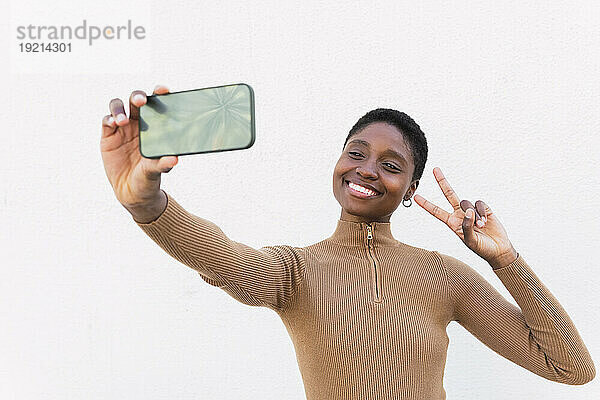 Glückliche Frau zeigt eine Geste des Friedenszeichens und macht ein Selfie vor der Wand