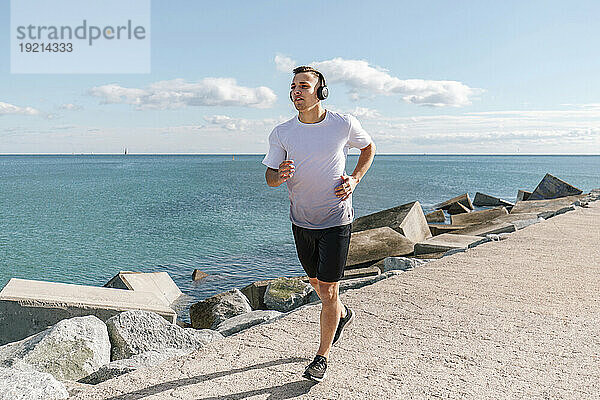 Junger Mann mit kabellosen Kopfhörern joggt am Pier am Meer