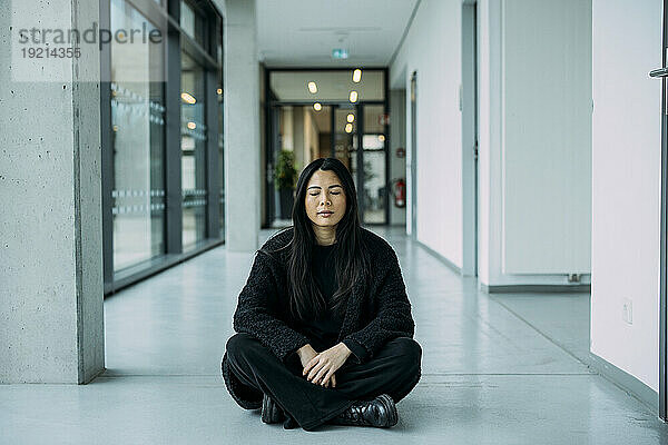 Geschäftsfrau sitzt mit gekreuzten Beinen und geschlossenen Augen auf dem Boden im Flur am Arbeitsplatz