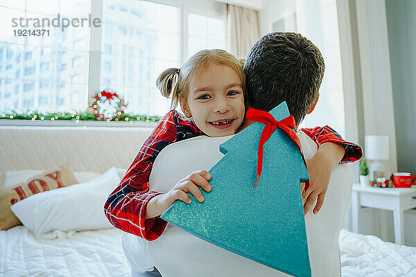 Glückliche Tochter hält Weihnachtsgeschenk in der Hand und umarmt Vater im Bett