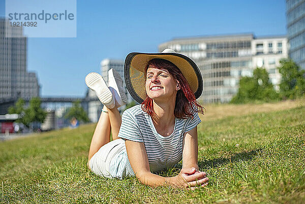 Lächelnde Frau mit Sonnenhut entspannt sich mit geschlossenen Augen im Gras