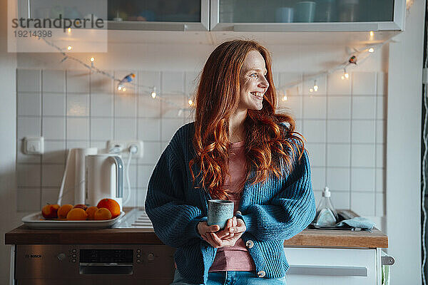 Glückliche rothaarige Frau steht mit einer Kaffeetasse zu Hause
