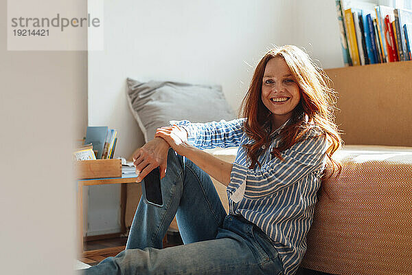 Happy redhead woman sitting with smart phone near bed at home