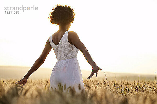 Junge Frau steht bei Sonnenuntergang mit ausgestreckten Armen im Weizenfeld