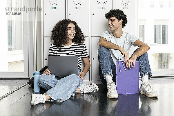 Lächelnde Studenten mit Laptop sitzen vor einem Schließfach in der Universität