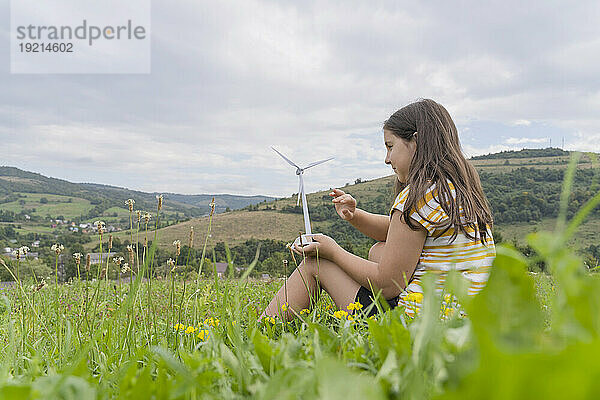 Mädchen mit Windturbinenmodell sitzt auf einer Wiese unter bewölktem Himmel