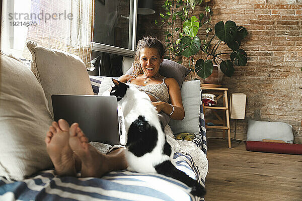 Lächelnde schwangere Frau mit Laptop und Katze sitzt zu Hause auf dem Sofa
