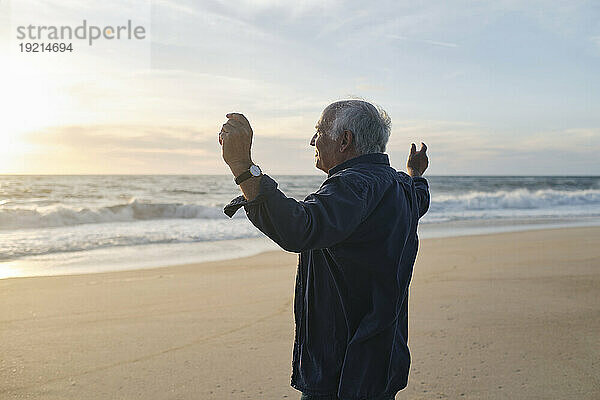 Älterer Mann mit erhobenen Armen steht am Strand