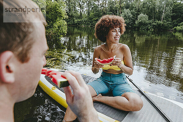 Nachdenkliche Frau isst mit Freundin Wassermelone auf dem Paddleboard im See