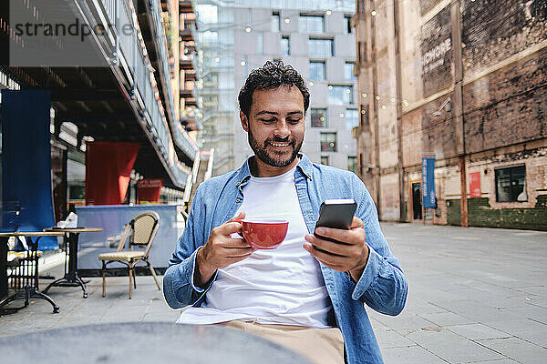 Lächelnder Mann hält Kaffeetasse in der Hand und benutzt Smartphone im Straßencafé