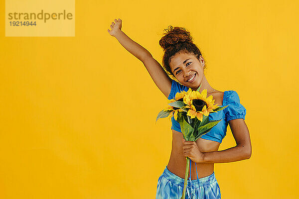Glückliche Frau mit erhobener Hand hält Sonnenblume vor gelbem Hintergrund