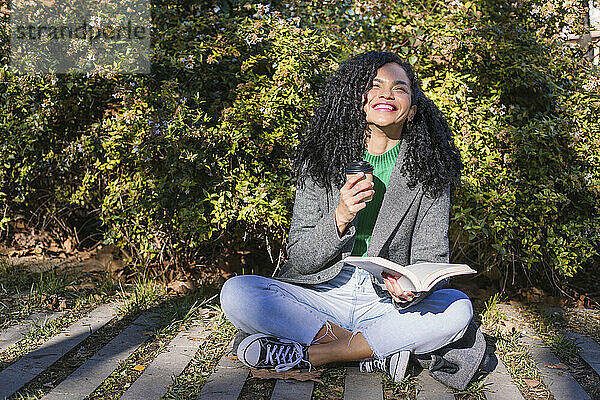 Happy woman with disposable coffee cup and book in park