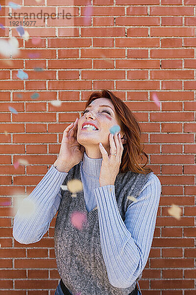 Happy woman with falling confetti in front of brick wall