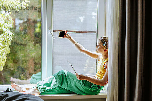 Girl taking selfie over smart phone sitting on window sill