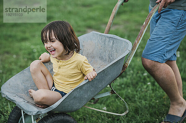 Lachender Junge sitzt in einer Schubkarre  die vom Vater geschoben wird