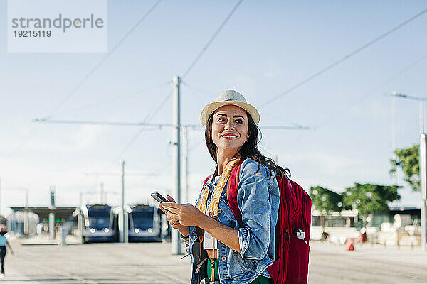 Glückliche junge Frau mit Mobiltelefon steht an einem sonnigen Tag
