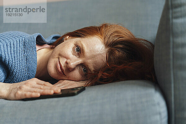Lächelnde Frau liegt mit Smartphone zu Hause auf grauem Sofa