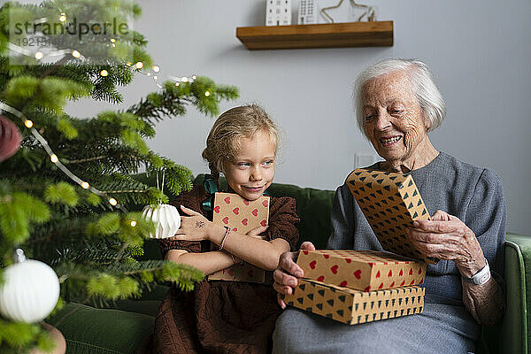 Glückliche Enkelin mit Großmutter  die zu Hause Geschenke in der Nähe des Weihnachtsbaums hält