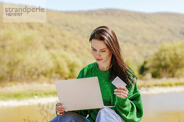 Lächelnde Frau nutzt Kreditkarte für Online-Einkäufe auf Laptop in der Nähe des Berges
