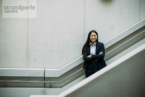 Smiling businesswoman with arms crossed leaning on staircase railing at workplace