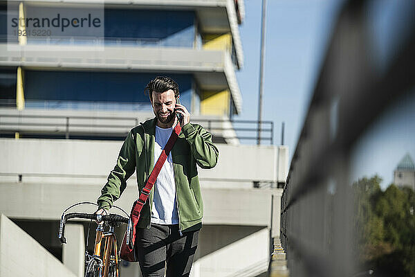 Lächelnder Mann  der mit dem Smartphone telefoniert und mit dem Fahrrad vor dem Gebäude läuft