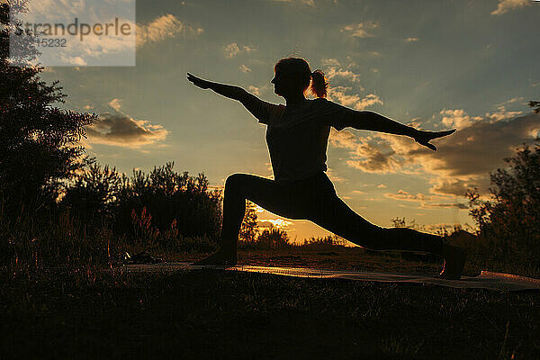 Silhouette einer Frau  die bei Sonnenuntergang Krieger-2-Yoga praktiziert