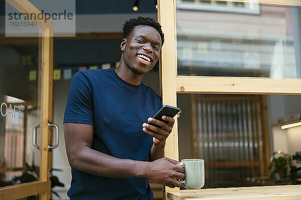 Fröhlicher junger Student steht mit Smartphone und Kaffeetasse da