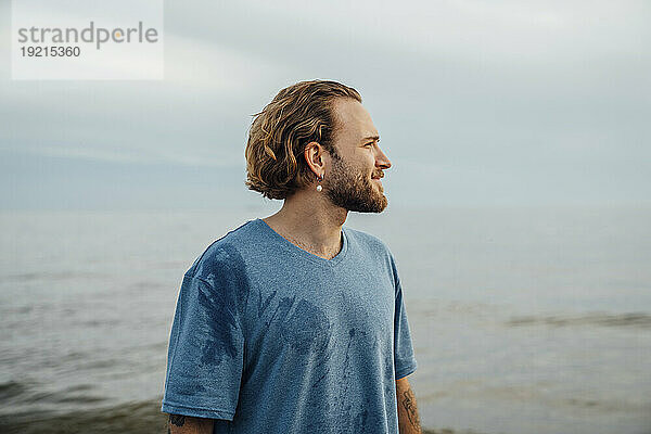 Lächelnder blonder Mann im T-Shirt in der Nähe des Meeres am Strand