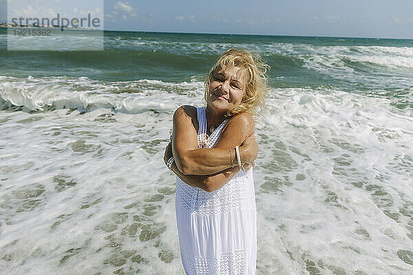 Lächelnde ältere Frau umarmt sich selbst und steht am Strand am Meer