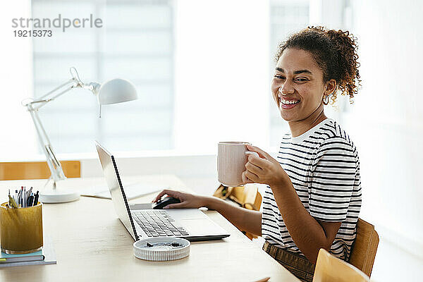 Fröhlicher junger Student hält Kaffeetasse in der Hand und sitzt mit Laptop am Schreibtisch