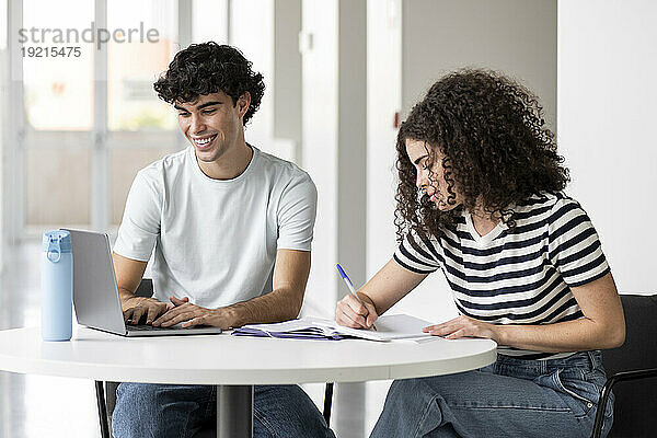 Lächelnde Studenten schreiben an Büchern und benutzen Laptop am Tisch in der Universität