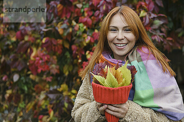 Glückliche rothaarige Frau hält Herbstblätter in Strickmütze im Park