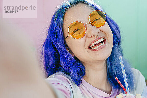 Fröhliche Hipster-Frau mit gefärbten blauen Haaren hält einen Drink in der Hand und macht ein Selfie im Café