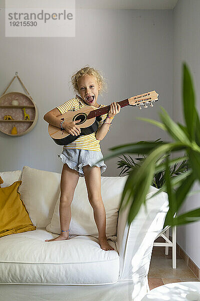 Glückliches Mädchen  das zu Hause auf der Couch im Wohnzimmer Gitarre spielt