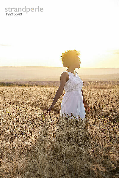 Junge Frau in weißem Kleid steht bei Sonnenuntergang im Weizenfeld