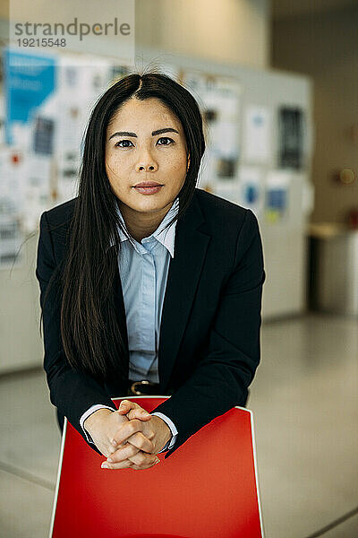 Confident businesswoman with hands clasped leaning on red chair at office