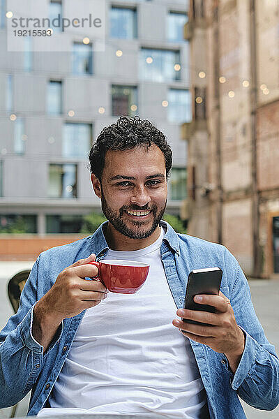 Lächelnder Mann sitzt mit Kaffeetasse und Smartphone vor Gebäuden