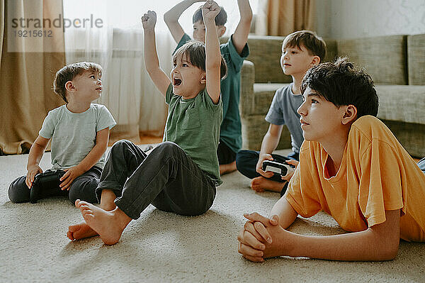 Glücklicher Junge spielt Videospiel mit Brüdern  die zu Hause auf dem Teppich jubeln