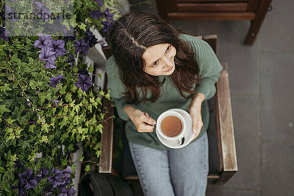 Nachdenkliche Frau hält Teetasse in der Hand und sitzt neben Pflanzen im Café