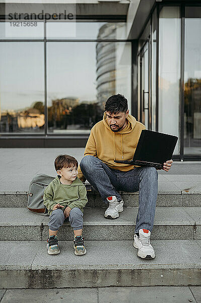 Vater hält Laptop in der Hand und sitzt mit Sohn auf Stufen