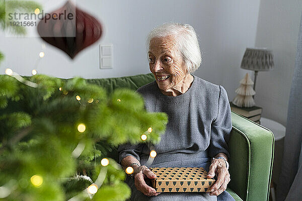 Glückliche ältere Frau sitzt mit Geschenkbox auf dem Sofa neben dem Weihnachtsbaum zu Hause