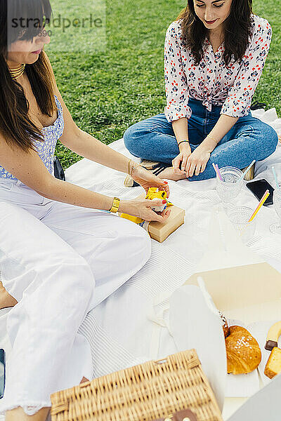 Lächelnde Frau mit Freundin öffnet Geschenkbox auf Picknickdecke