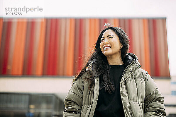 Lächelnde schöne Geschäftsfrau in gepolsterter Jacke vor dem Bürogebäude