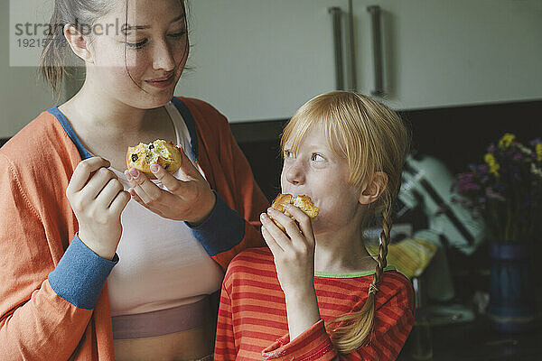 Blond girl eating muffins with elder sister in kitchen at home