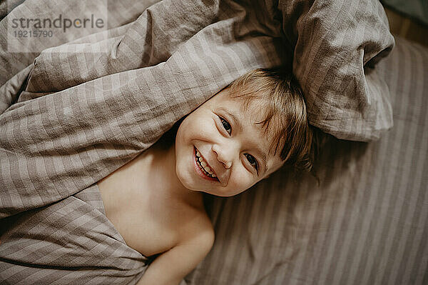 Lächelnder Junge liegt zu Hause mit Decke auf dem Bett