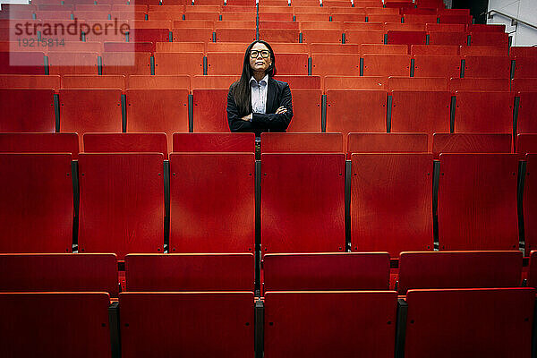 Confident businesswoman with arms crossed amidst red seats in convention center