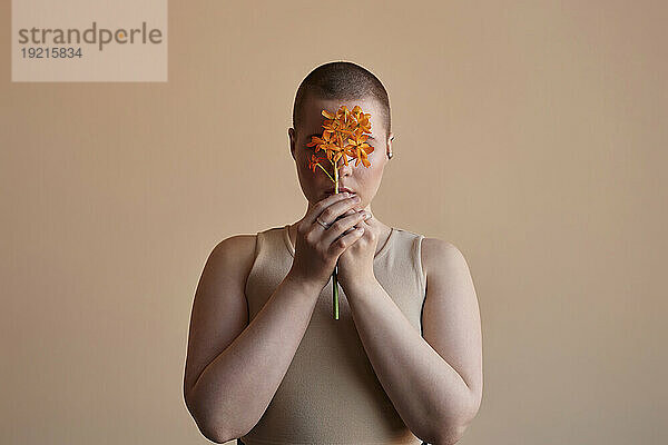 Frau bedeckt Gesicht mit orangefarbener Blume vor beigem Hintergrund