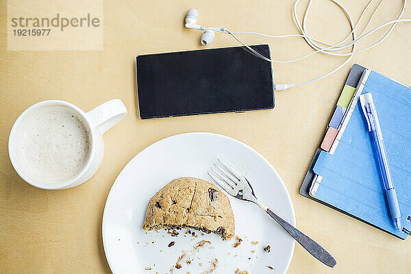 Kaffeetasse mit Keksscheibe auf dem Teller neben Smartphone und Milchprodukten am Tisch