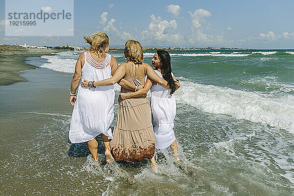 Friends with arms around walking together on shore at beach