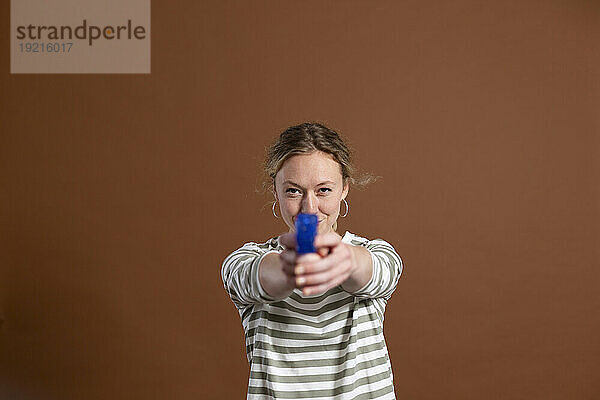 Blonde Frau hält Spielzeugpistole vor braunem Hintergrund
